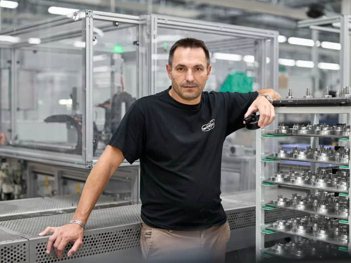 Foto: Gesco: Mann in schwarzen Shirt in Produktionshalle, Paletten von Kleinteilen neben ihm